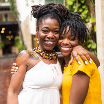 Portrait photo outdoors of mother and daughter smiling at camera with arms around each other's shoulders.