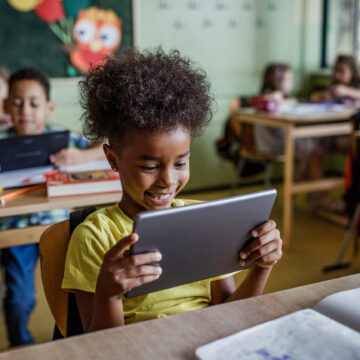Black elementary student smiling in classroom, sitting at a desk and looking at a handheld electronic tablet.
